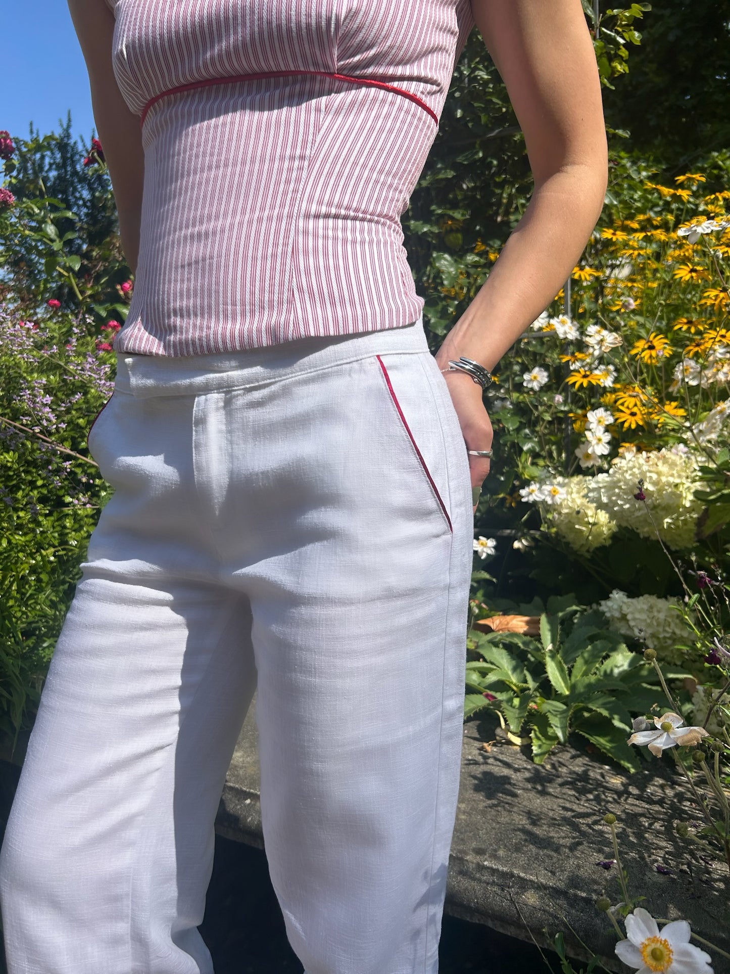 Woman wearing a pink top and white linen pants with pink piping, standing in a garden.