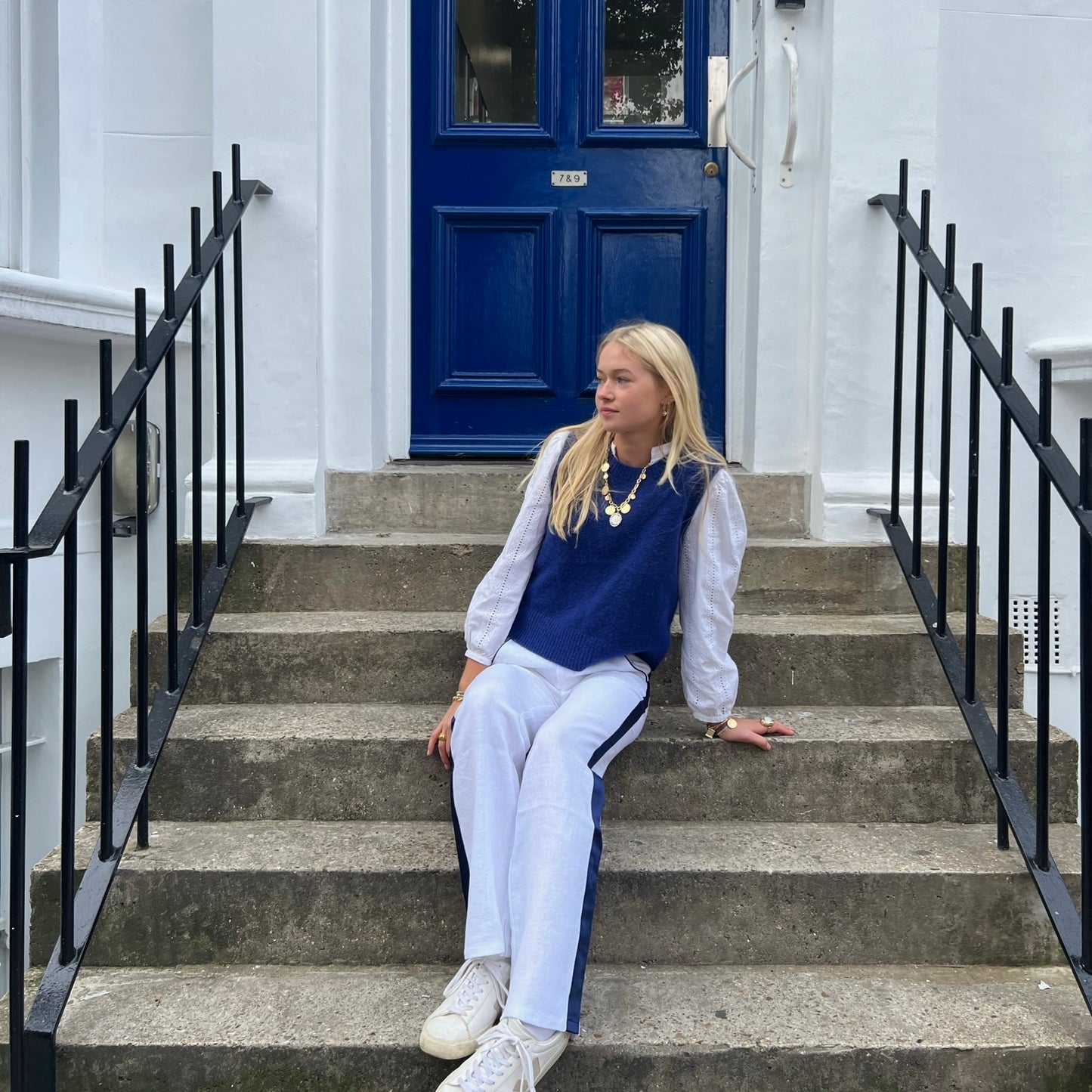 Woman wearing white linen trousers with a navy stripe down the side, sitting outside a blue doorway