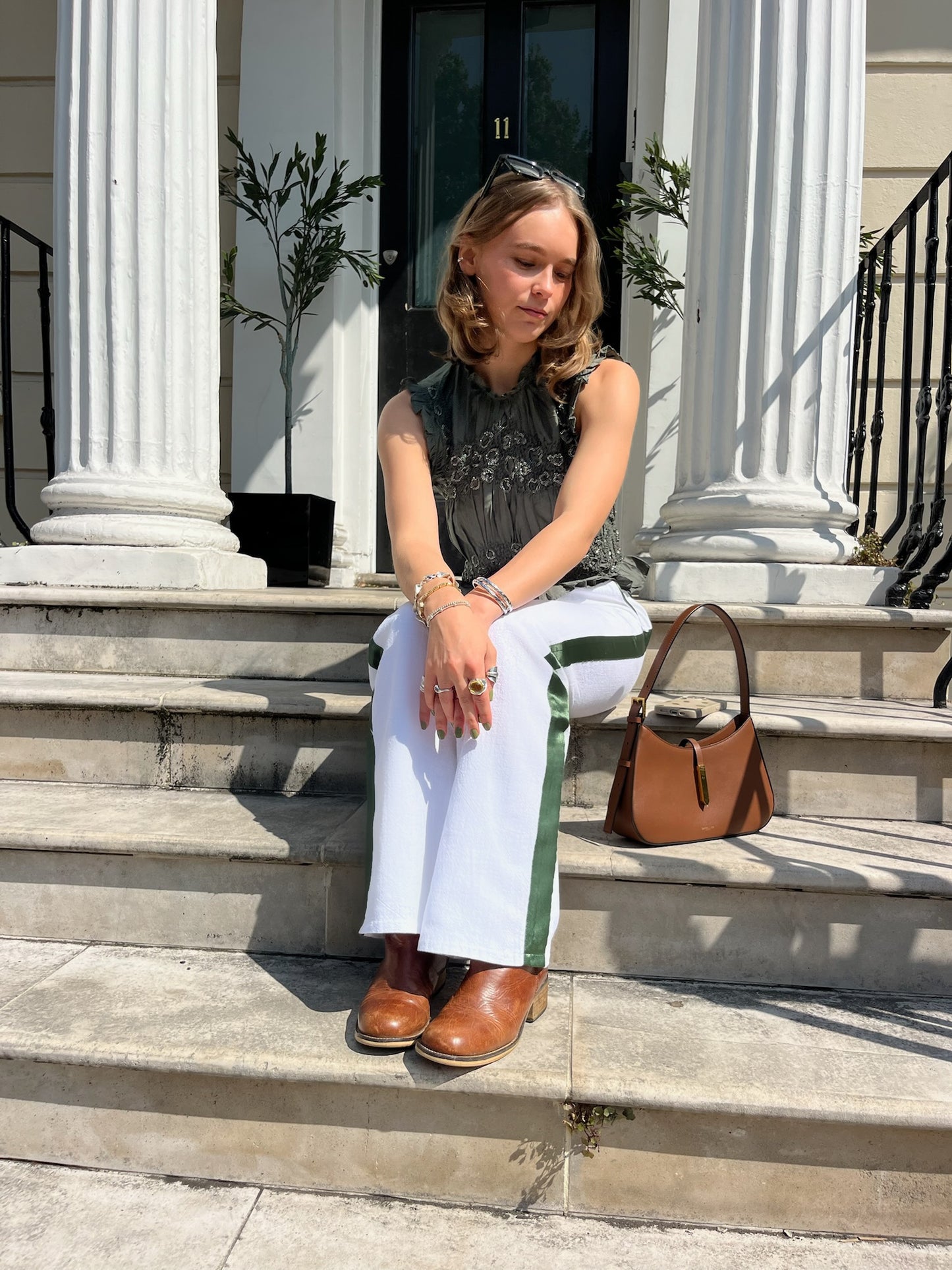 Woman sitting on steps wearing a green top, white linen trousers with a green stripe and brown shoes, with a brown handbag.