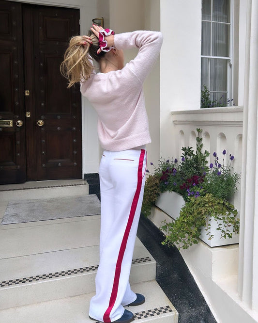 A young woman stands in a doorway entrance in London, wearing Moony linen trousers in berry color. She is holding her hair with both hands. The setting and her attire exude a blend of urban chic and sustainable fashion