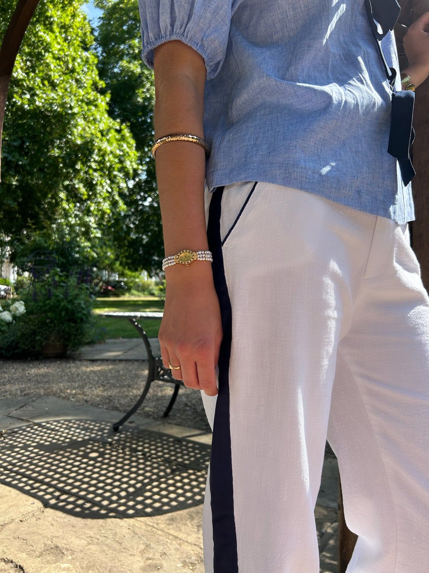 Woman wearing a blue shirt and white linen trousers with a navy detail and a park setting in the background