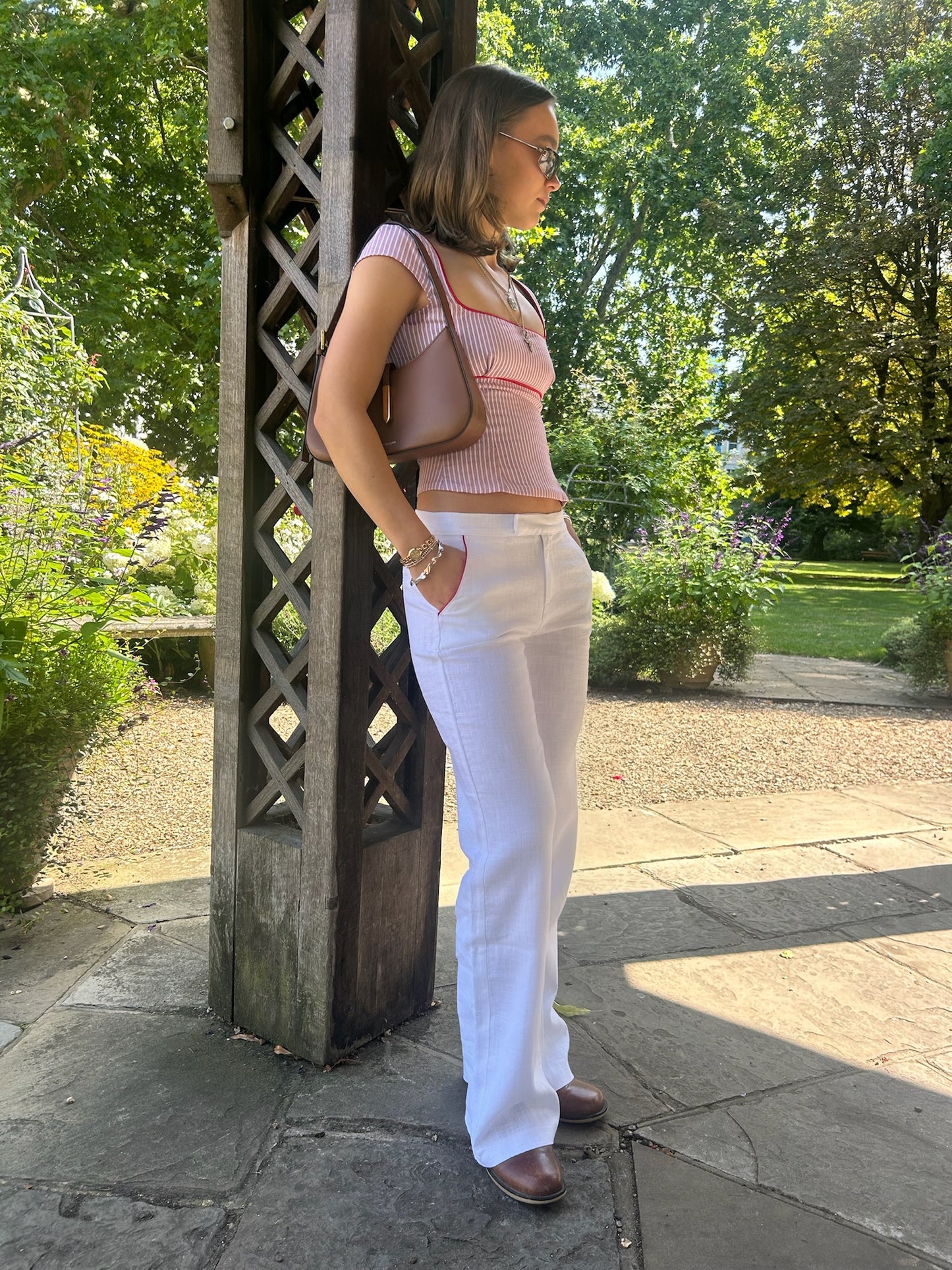 Woman in a garden wearing a pink top and white linen trousers, standing next to a wooden lattice structure.