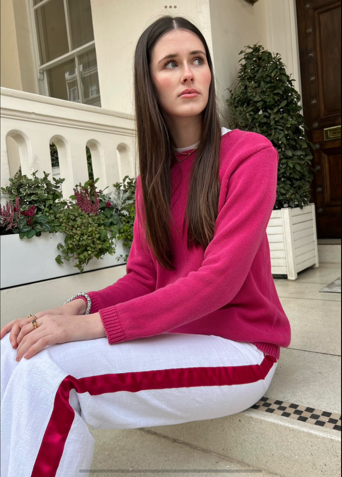 Woman sitting on steps wearing a pink jumper and white linen trousers with a pink stripe down the side