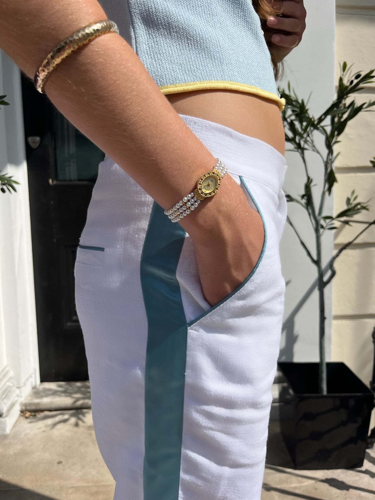 A woman wearing white linen trousers with teal detailing down the side and on the pockets, a gold watch, and bracelet.